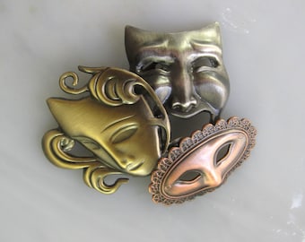 May include: Three metal masks in a cluster. The masks are in the style of traditional theater masks. One mask is silver and has a sad expression. Another mask is gold and has a neutral expression. The third mask is copper and has a masquerade design.
