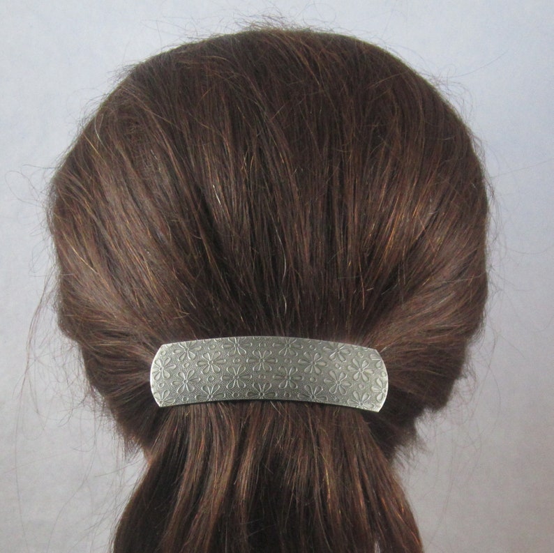 Puede incluir: Una pinza para el pelo de plata con un patr&oacute;n floral. La pinza tiene forma de rect&aacute;ngulo y se utiliza para sujetar una coleta.