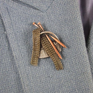 Pode incluir: Broche vintage com tema de tricô. O alfinete tem uma cesta, agulhas de tricô e um design semelhante a uma corrente em tons de prata, cobre e ouro. O broche é preso a um blazer azul.
