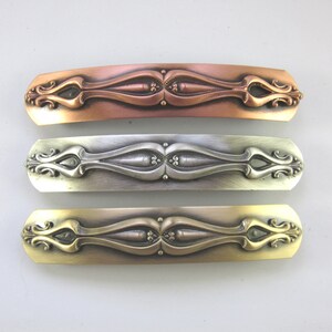 K&ouml;nnte beinhalten: Drei dekorative Haarklammern mit einem verzierten Design. Die Klammern sind aus Metall und in Kupfer, Silber und Gold ausgef&uuml;hrt.