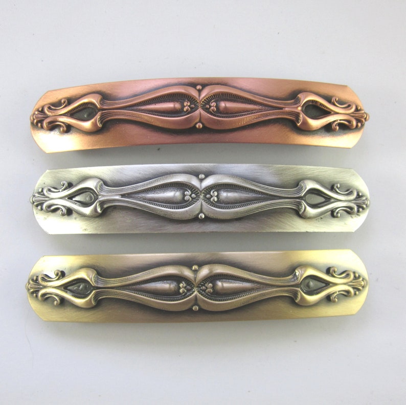 K&ouml;nnte beinhalten: Drei dekorative Haarklammern mit einem verzierten Design. Die Klammern sind aus Metall und in Kupfer, Silber und Gold ausgef&uuml;hrt.