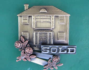 May include: A gold-toned metal house figurine with a 'SOLD' sign and rose accents. The house has a traditional design with a front door and windows.