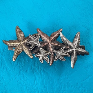 May include: A hair clip featuring five metal starfish in a cluster. The starfish are in shades of gold, silver, and copper.