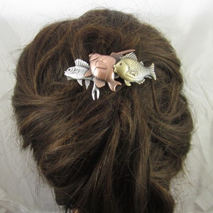 Puede incluir: Una pinza para el pelo de plata, oro y cobre con tres dise&ntilde;os de peces. Los peces son estilizados y tienen un aspecto ligeramente abstracto. La pinza es un accesorio decorativo para el cabello.