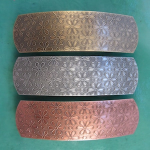 Puede incluir: Tres pinzas para el pelo de metal con un patr&oacute;n de margarita. La pinza superior es dorada, la pinza del medio es plateada y la pinza inferior es de cobre.