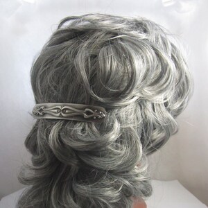 K&ouml;nnte beinhalten: Eine silberne Haarklammer mit einem verzierten Design. Die Klammer hat die Form eines Rechtecks mit einer abgerundeten Ober- und Unterseite. Das Design zeigt eine Reihe von Wirbeln und Punkten.