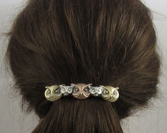 May include: A silver, gold, and copper owl hair clip with five owl heads. The owls are facing forward with their eyes open.