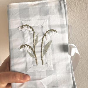 May include: A rectangular fabric book cover featuring a light blue and white gingham pattern. The center displays an embroidered design of white lily of the valley flowers with green stems and leaves, framed by a white rectangular stitched border.
