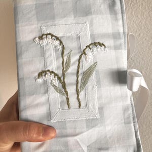 May include: A rectangular fabric book cover featuring a light blue and white gingham check pattern. The center displays an embroidered design of two lily of the valley stems with white bell-shaped flowers and green leaves, framed by a stitched rectangle.