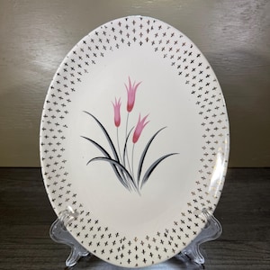 May include: An oval-shaped white ceramic platter featuring a central graphic of three pink tulip-like flowers with long, curved gray leaves. The border of the platter is decorated with a repeating pattern of small, metallic gold cross symbols.