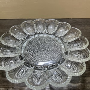 May include: A clear glass egg serving platter featuring a circular center with a textured hobnail pattern surrounded by twelve individual oval-shaped indentations, each with a scalloped edge and a detailed embossed shell-like design.