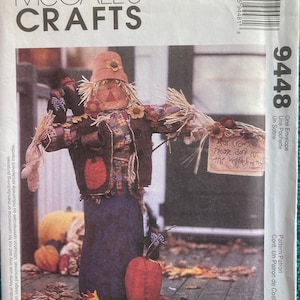 May include: McCall's Crafts pattern for a scarecrow. The scarecrow has a straw hat, a patchwork jacket, and a blue skirt. It holds a sign that reads "Dear Crows, Please don't pat the veggies". Pumpkins and fall leaves are in the background.