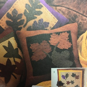 May include: Three brown, yellow, and purple throw pillows with various leaf designs. The pillows are on a brown leather couch. A book and glasses are on a table in the foreground. A wall hanging with a similar leaf design is in the background.