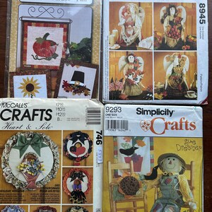 May include: A collection of vintage craft pattern packets. The packets feature designs for seasonal decorations, including a calendar, angel figures, wreaths, and a scarecrow. The packets are in various sizes and colors, with text and illustrations.
