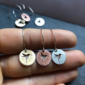 Puede incluir: Tres pares de pendientes de aro de plata con un diseño de colibrí recortado. Los pendientes se muestran en tres acabados metálicos diferentes: oro, cobre y plata.