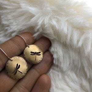 Puede incluir: Un par de pendientes de color dorado con un diseño de libélula. Los pendientes cuelgan de una cadena plateada y están sobre una superficie blanca y peluda.