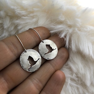 Puede incluir: Un par de pendientes de plata con un diseño recortado de un pájaro en vuelo. Los pendientes tienen una textura martillada y están suspendidos de un aro simple.