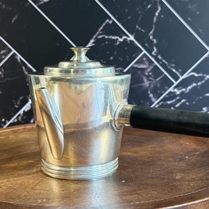 May include: A silver-toned metal teapot with a cylindrical body, a short spout, and a flat lid with a small knob. The teapot features a horizontal black handle extending from the side and a series of horizontal ridges around the base.