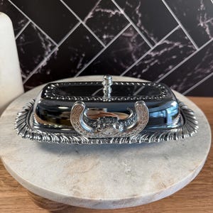 May include: A rectangular silver-toned butter dish with a polished reflective lid and a decorative handle. The base features an ornate scalloped edge with intricate embossed detailing, and the lid is topped with a small, rounded knob finial.