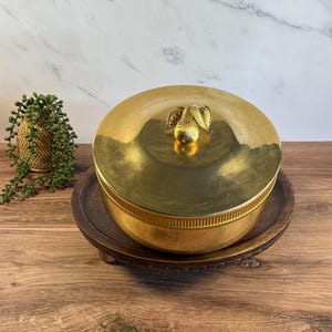 May include: A round, polished brass container with a lid featuring a decorative pear-shaped handle. The container has a smooth, reflective surface and a textured, ribbed band around the middle section of the base, providing a metallic finish.