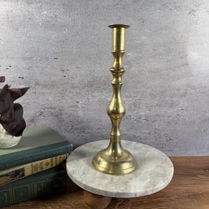 May include: A tall, polished brass candlestick holder with a slender, multi-tiered stem and a wide, circular base. The metal surface has a warm, golden metallic sheen, featuring a smooth, reflective finish that catches the light along its curves.