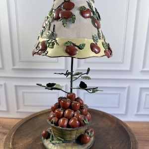 May include: A table lamp featuring a mesh shade decorated with metal red apples and green leaves. The base is a basket overflowing with red apples, supported by a thin metal stem with additional leaf accents. The lamp has a cream-colored trim.