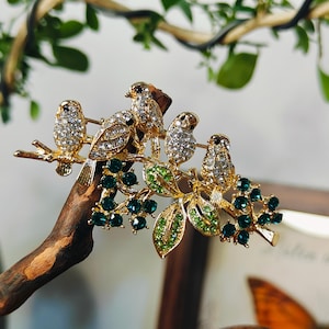 Peut inclure: Une broche en métal doré représentant quatre oiseaux perchés sur une branche. Les oiseaux sont incrustés de cristaux clairs. La branche est ornée de grappes de cristaux ronds vert foncé et de feuilles en cristaux vert clair.