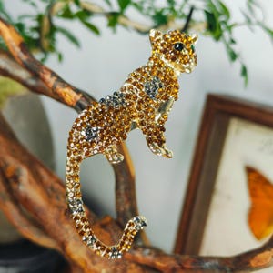 Peut inclure: Une broche en métal doré en forme de léopard, entièrement incrustée de strass ambre et gris foncé. La figure féline est représentée dans une pose debout avec une longue queue courbée, dotée d'un seul œil en strass vert foncé.