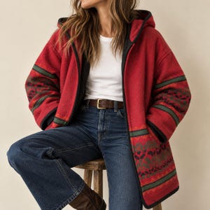 Puede incluir: Una chaqueta roja con capucha que presenta una banda estampada en verde oscuro y negro en las mangas y el dobladillo, sobre una camiseta blanca y vaqueros azules con un cinturón de cuero marrón. La chaqueta tiene bordes negros y capucha.