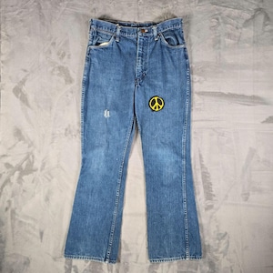 May include: Blue denim jeans featuring a high-waisted cut, a classic five-pocket design, and a yellow and black peace sign patch stitched onto the upper right thigh area. The fabric shows a medium wash with subtle fading and a small distressed mark.