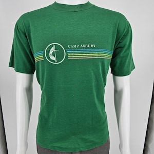 May include: A short-sleeved green t-shirt featuring a white circular logo with a cross and flame emblem. To the right of the logo, the text CAMP ASBURY is printed above a series of horizontal lines in shades of blue, green, and yellow.