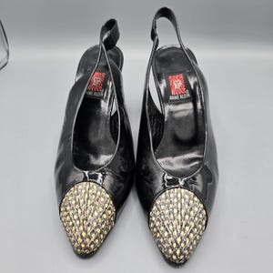 May include: A pair of black patent leather slingback heels with a pointed toe featuring a textured, yellow and black snakeskin-patterned cap. The interior footbed displays a red and white square logo with a lion graphic and the text ANNE KLEIN.