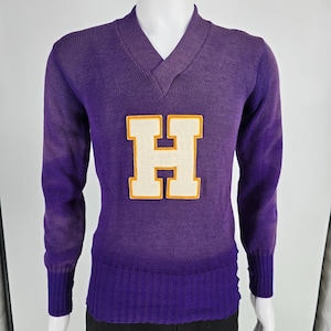Peut inclure: Un pull en tricot violet à manches longues avec un col en V et une grande lettre H appliquée au centre. La lettre H est de couleur crème avec une bordure orange épaisse. Le pull présente des poignets côtelés et un large ourlet côtelé.