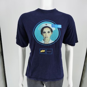 May include: A dark blue short-sleeve t-shirt featuring a graphic of a nurse in a white cap with a finger to her lips. The design includes a light blue circle, the text "MIXED BY SCHWEPPES", and a small yellow Schweppes logo on the front chest.