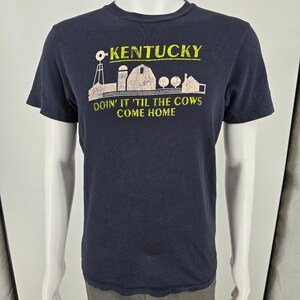 May include: A dark navy blue short-sleeve t-shirt featuring a graphic print of a farm scene with a windmill, barn, and silos. The text reads KENTUCKY in yellow and DOIN' IT 'TIL THE COWS COME HOME in yellow below the farm illustration.