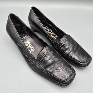 Puede incluir: Un par de zapatos de tacón de cuero negro con tacón bajo y punta cuadrada. El exterior presenta un patrón circular repetitivo en relieve en toda la superficie. La plantilla interior es negra y muestra el nombre de la marca Salvatore Ferragamo en oro.