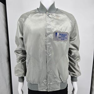 May include: A light grey satin bomber jacket with a ribbed collar, cuffs, and hem. The jacket features a front snap-button closure and a rectangular patch on the left chest that reads WELLS ALUMINUM MONETT/CASSVILLE DIVISION in blue text.