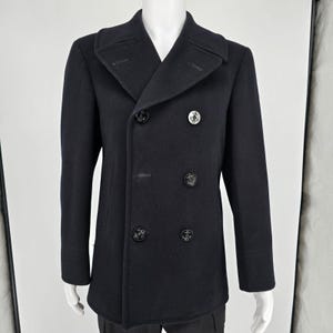 May include: A dark navy blue double-breasted pea coat featuring wide lapels and six black buttons embossed with anchor motifs. The coat has a structured silhouette with long sleeves and vertical side pockets, displayed on a mannequin.