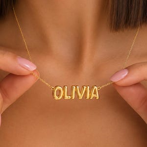 May include: A gold-toned necklace featuring a pendant with the name OLIVIA written in bold, rounded, three-dimensional capital letters. The pendant is attached to a delicate, thin gold-toned chain that rests around the neck area.