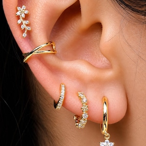 May include: A set of gold-toned earrings including a floral climber, a crisscross ear cuff, two small pavé-set huggie hoops, and a single hoop earring featuring a dangling six-petaled flower charm encrusted with clear white stones.