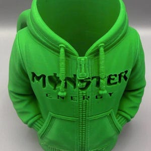 May include: A bright green mug shaped like a hooded sweatshirt with a zip detail and drawstrings. The front features the embossed text "MONSTER ENERGY" across the chest area, mimicking the design of a zip-up hoodie with ribbed cuffs and hem.