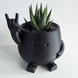Peut inclure: Un pot de fleurs noir mat en forme de personnage rond avec deux petits pieds et deux bras. Un bras est levé dans un geste de la main rock-and-roll, tandis que l'autre repose sur le côté. Le visage présente deux yeux circulaires et un petit sourire. Une plante succulente verte est à l'intérieur.