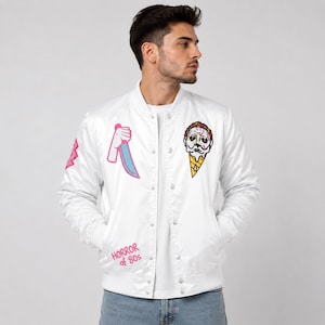 Puede incluir: Una chaqueta bomber blanca con un gráfico de una mano sosteniendo un cuchillo azul, un cono de helado derritiéndose con una cara y el texto rosa "HORROR of 80s" impreso en la parte delantera. La chaqueta tiene cierre de botones a presión.