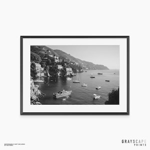 May include: A black and white framed landscape photograph depicting a coastal village nestled on a hillside overlooking a bay filled with various boats and yachts. The scene features rocky cliffs, Mediterranean-style buildings, and calm water.