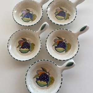 Pode incluir: Um conjunto de cinco tigelas de cerâmica em tom esbranquiçado com pegas, cada uma com um cacho de uvas roxas e folhas verdes pintadas à mão no centro. As bordas têm uma linha tracejada verde-escura e cada pega apresenta uma folha verde.