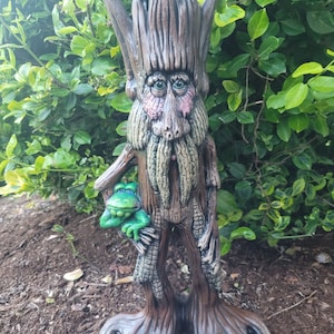 May include: A whimsical tree-like figurine with a human-like face, wearing a long beard and holding a small green frog. The figurine is made of brown and green ceramic and has a textured surface.