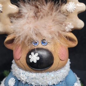 Puede incluir: Una figurita de reno de cer&aacute;mica con pelaje marr&oacute;n, ojos azules y nariz negra. El reno lleva un su&eacute;ter azul con copos de nieve blancos y un ribete de piel blanca. El reno tiene un copo de nieve blanco en la nariz.