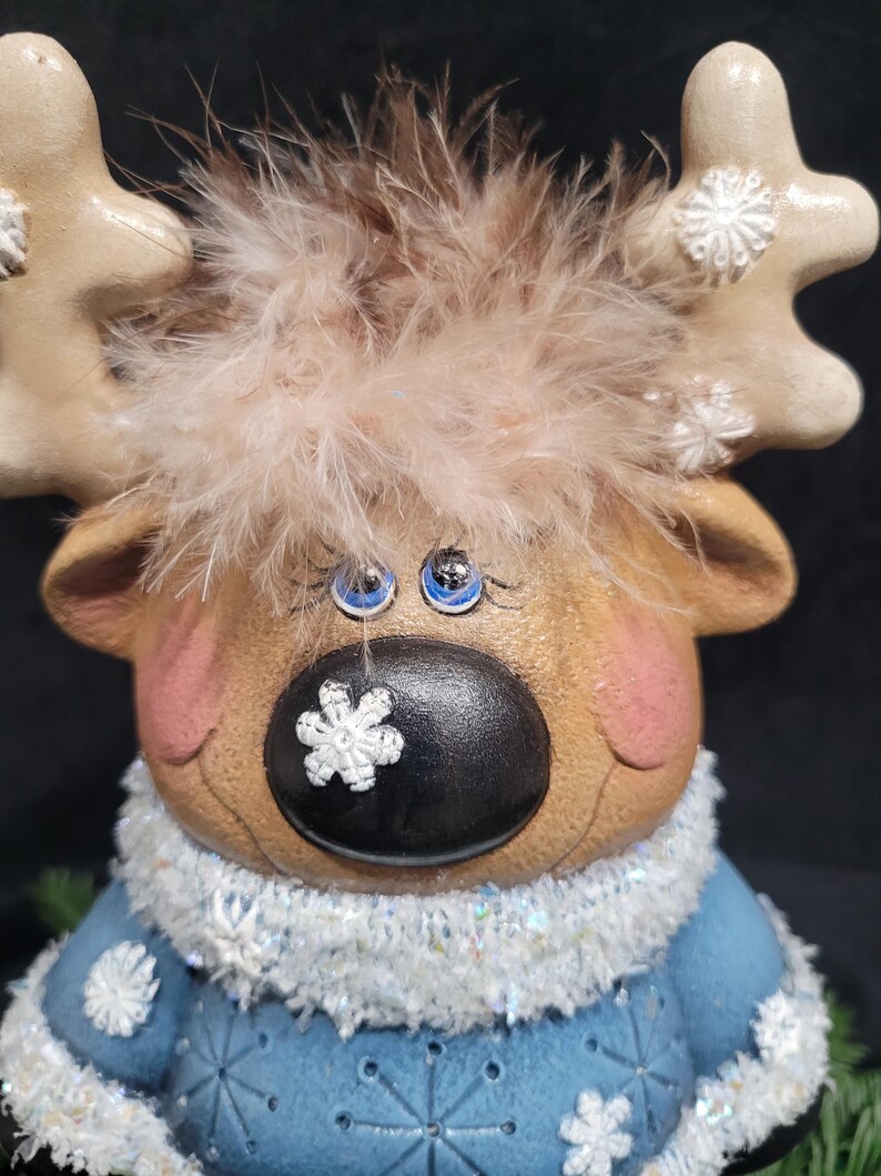 Puede incluir: Una figurita de reno de cer&aacute;mica con pelaje marr&oacute;n, ojos azules y nariz negra. El reno lleva un su&eacute;ter azul con copos de nieve blancos y un ribete de piel blanca. El reno tiene un copo de nieve blanco en la nariz.