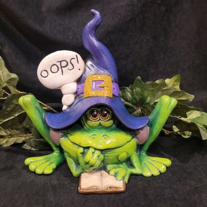 May include: A green frog wearing a purple and blue witch hat with a gold buckle and a white speech bubble that says "OOPS!" The frog is holding a book in its hands.