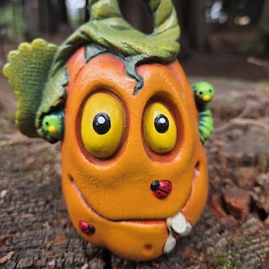 May include: A sculpted orange pumpkin figure with large yellow eyes, a green stem handle, and a green leaf. The pumpkin features a small green caterpillar on the side, two red ladybugs on its surface, and a white tooth protruding from its mouth.